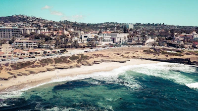 A coastal town with a sandy beach and rocky formations. Waves crash along the shoreline, where people can be seen enjoying the sun and water. In the background, there is a bustling town with numerous buildings and palm trees lining the streets.