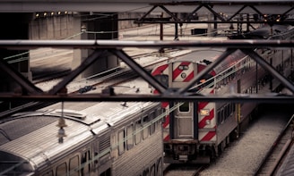 An urban scene with a complex network of train tracks and two metallic trains. The image captures a perspective from an overpass, showcasing the sleek design of the train cars beneath the framework of the bridge. The area appears industrial, with concrete walls and overhead lights.