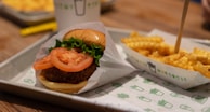 A close-up of a fast food meal featuring a burger with a breaded patty, topped with fresh lettuce and tomato slices, served on a tray lined with branded paper. Next to the burger is a portion of crinkle-cut fries topped with cheese sauce, placed in a branded carton.