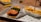 A close-up of a fast food meal featuring a burger with a breaded patty, topped with fresh lettuce and tomato slices, served on a tray lined with branded paper. Next to the burger is a portion of crinkle-cut fries topped with cheese sauce, placed in a branded carton.