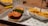 A close-up of a fast food meal featuring a burger with a breaded patty, topped with fresh lettuce and tomato slices, served on a tray lined with branded paper. Next to the burger is a portion of crinkle-cut fries topped with cheese sauce, placed in a branded carton.