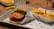 A close-up of a fast food meal featuring a burger with a breaded patty, topped with fresh lettuce and tomato slices, served on a tray lined with branded paper. Next to the burger is a portion of crinkle-cut fries topped with cheese sauce, placed in a branded carton.