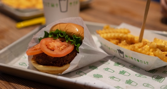 A close-up of a fast food meal featuring a burger with a breaded patty, topped with fresh lettuce and tomato slices, served on a tray lined with branded paper. Next to the burger is a portion of crinkle-cut fries topped with cheese sauce, placed in a branded carton.