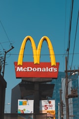 A prominent McDonald's sign featuring the iconic golden arches and the company name is displayed against a clear blue sky. Power lines and poles are visible in the foreground, and the lower portion of the image shows advertisements with promotional offers.