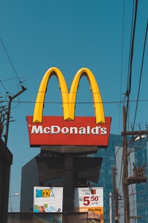A prominent McDonald's sign featuring the iconic golden arches and the company name is displayed against a clear blue sky. Power lines and poles are visible in the foreground, and the lower portion of the image shows advertisements with promotional offers.