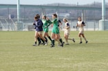 A group of female soccer players jogging on a grass field. Some are wearing green uniforms while others are in beige. In the background, there is a bridge and a chain-link fence. The atmosphere is casual and seems like a practice or a break during a game.