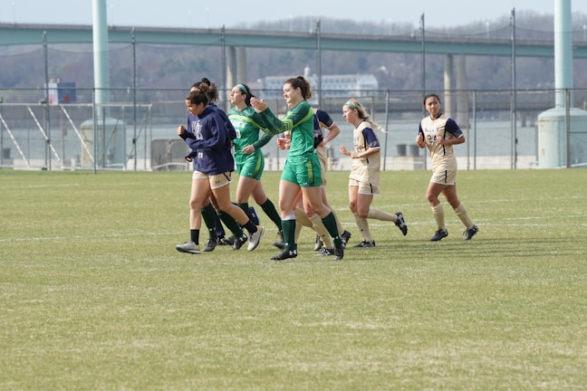 A group of female soccer players jogging on a grass field. Some are wearing green uniforms while others are in beige. In the background, there is a bridge and a chain-link fence. The atmosphere is casual and seems like a practice or a break during a game.