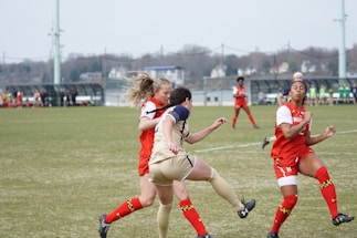 A women's soccer match is taking place with players intensely competing for the ball. The players are wearing red and white uniforms from one team and beige uniforms from the other. A green grassy field and blurred spectators in benches are visible in the background.