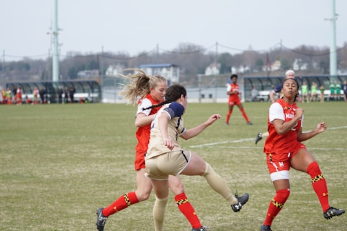 A women's soccer match is taking place with players intensely competing for the ball. The players are wearing red and white uniforms from one team and beige uniforms from the other. A green grassy field and blurred spectators in benches are visible in the background.