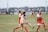 A women's soccer match is taking place with players intensely competing for the ball. The players are wearing red and white uniforms from one team and beige uniforms from the other. A green grassy field and blurred spectators in benches are visible in the background.