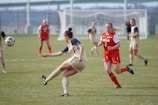 A women's soccer match is underway on a grassy field. One player, in a tan and navy uniform, is kicking the ball while another player in a red and white Maryland jersey is running toward her. Other players are scattered across the field, and a goalpost is visible in the background.