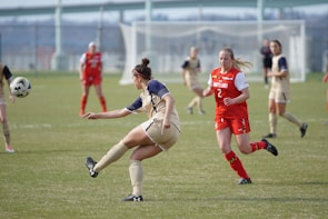 A women's soccer match is underway on a grassy field. One player, in a tan and navy uniform, is kicking the ball while another player in a red and white Maryland jersey is running toward her. Other players are scattered across the field, and a goalpost is visible in the background.