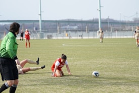 A soccer match is taking place on a grassy field. One player in a red and white uniform is kneeling on the ground near a soccer ball, appearing to recover from a fall. Another player in a beige uniform is also on the ground. A referee in a green and black uniform is observing the scene. There are several players in the background, some running and others standing. A blurred cityscape and bridge are visible in the distance.