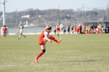 A soccer player in a red jersey and shorts, with a black headband, is captured mid-air as they perform a dynamic kick. The soccer ball is close to their foot, indicating an intense moment of action. In the background, other players and people are visible near the sideline, with goalposts and a net surrounding the field. The scene is set on a green grass field with a hazy view of buildings and trees in the distance.