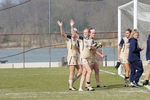 A group of female soccer players is gathered near a goalpost on a grassy field. They are wearing beige uniforms with the letter 'N' on the front. Two of the players are waving happily. A few players are holding cleats in their hands. The background includes a soccer net and a partly blurred scenic view with trees.