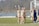 A group of female soccer players is gathered near a goalpost on a grassy field. They are wearing beige uniforms with the letter 'N' on the front. Two of the players are waving happily. A few players are holding cleats in their hands. The background includes a soccer net and a partly blurred scenic view with trees.