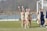 A group of female soccer players is gathered near a goalpost on a grassy field. They are wearing beige uniforms with the letter 'N' on the front. Two of the players are waving happily. A few players are holding cleats in their hands. The background includes a soccer net and a partly blurred scenic view with trees.