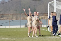 A group of female soccer players is gathered near a goalpost on a grassy field. They are wearing beige uniforms with the letter 'N' on the front. Two of the players are waving happily. A few players are holding cleats in their hands. The background includes a soccer net and a partly blurred scenic view with trees.