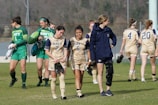A group of female soccer players wearing navy and green uniforms are walking together on a grass field. Some players are carrying cleats, and one has a knee brace. The goalpost and trees are visible in the background.