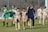 A group of female soccer players wearing navy and green uniforms are walking together on a grass field. Some players are carrying cleats, and one has a knee brace. The goalpost and trees are visible in the background.