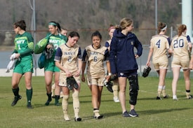 A group of female soccer players wearing navy and green uniforms are walking together on a grass field. Some players are carrying cleats, and one has a knee brace. The goalpost and trees are visible in the background.