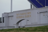 The image displays the entrance of the Navy-Marine Corps Memorial Stadium with large, prominent lettering. The structure is made of pale stone and features inscriptions for different graduating classes. Part of the stadium's seating area with blue seats is visible in the background.