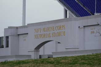 The image displays the entrance of the Navy-Marine Corps Memorial Stadium with large, prominent lettering. The structure is made of pale stone and features inscriptions for different graduating classes. Part of the stadium's seating area with blue seats is visible in the background.