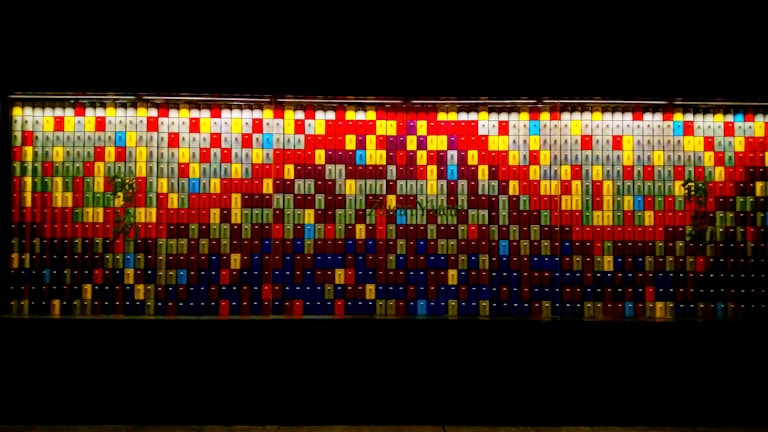 A vibrant wall display made up of numerous rectangular tiles arranged in a mosaic pattern, featuring a variety of colors including red, blue, yellow, and green. The tiles create an abstract, pixelated artwork with a complex design.