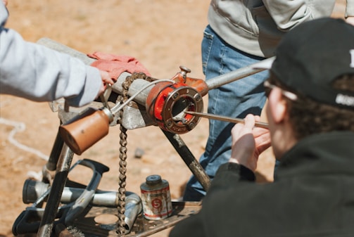 Several individuals are engaged in a hands-on activity focusing on mechanical equipment. The setup includes a metal pipe clamped into a red pipe threading tool, which is mounted on a tripod. Nearby, various tools and a canister are scattered on the ground. The background features an outdoor setting with dry, brown soil.