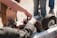 Hands are working on assembling or repairing a small wheeled device, possibly a robot or machine, with visible gears and metal components. Several wheels with thick treads are attached to a metal frame. A group of people stands around, suggesting teamwork or collaboration.