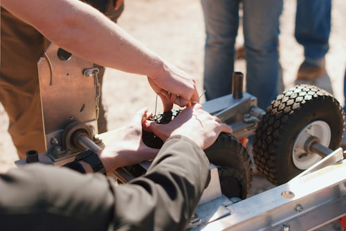 Hands are working on assembling or repairing a small wheeled device, possibly a robot or machine, with visible gears and metal components. Several wheels with thick treads are attached to a metal frame. A group of people stands around, suggesting teamwork or collaboration.