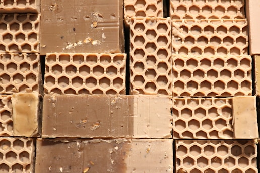 Several blocks of honeycomb-patterned beeswax are stacked closely together, displaying a textured surface with visible hexagonal impressions. The beeswax appears slightly rough, with some minor debris scattered throughout.