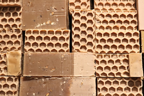 Several blocks of honeycomb-patterned beeswax are stacked closely together, displaying a textured surface with visible hexagonal impressions. The beeswax appears slightly rough, with some minor debris scattered throughout.