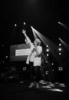 A singer is performing on stage with one hand raised while holding a microphone in the other. Bright stage lights create dramatic beams, and a screen in the background displays lyrics. A band can be seen in the dimly lit area behind the singer.