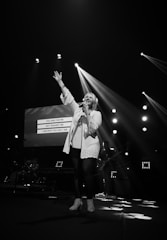 A singer is performing on stage with one hand raised while holding a microphone in the other. Bright stage lights create dramatic beams, and a screen in the background displays lyrics. A band can be seen in the dimly lit area behind the singer.