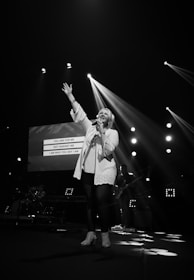 A singer is performing on stage with one hand raised while holding a microphone in the other. Bright stage lights create dramatic beams, and a screen in the background displays lyrics. A band can be seen in the dimly lit area behind the singer.