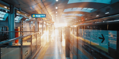 A modern train station with an empty platform is lit by bright sunlight streaming in from large windows overhead. The platform is clean and sleek, with a train on one side reflecting the light. Signage in a foreign language is visible above, along with digital clocks displaying the time.