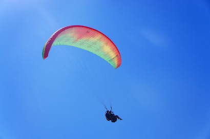 A paraglider with a colorful canopy is soaring through a clear, bright blue sky. The canopy features a gradient of green to red colors. The paraglider is suspended beneath, connected by lines, creating an image of freedom and adventure.