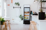 A minimalist and clean interior of a small cafe or shop. There is a refrigerator filled with various fruits like apples, oranges, and pears. Above the refrigerator, a sign reads 'GENUINE'. To the left of the refrigerator is a hanging white shirt and a shelf with a plant. The room has a few potted plants and wooden stools around a white countertop. Natural light streams in from windows, giving a bright and airy feel to the space.