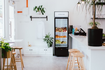 A minimalist and clean interior of a small cafe or shop. There is a refrigerator filled with various fruits like apples, oranges, and pears. Above the refrigerator, a sign reads 'GENUINE'. To the left of the refrigerator is a hanging white shirt and a shelf with a plant. The room has a few potted plants and wooden stools around a white countertop. Natural light streams in from windows, giving a bright and airy feel to the space.