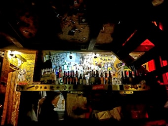 A dimly lit bar with a variety of bottles displayed on shelves. The walls are covered with graffiti and posters, creating an eclectic and rustic atmosphere. Glasses hang upside down above the bar area.
