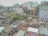 An urban skyline with a mix of residential and commercial buildings in varying states of repair. Some buildings are brightly colored, while others have a more aged appearance. A small number of trees and greenery are interspersed among the buildings, suggesting a densely populated area. Rooftops show a variety of materials and some are used for drying clothes. The area appears to be bustling with activity, indicated by people and objects scattered around.