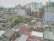 An urban skyline with a mix of residential and commercial buildings in varying states of repair. Some buildings are brightly colored, while others have a more aged appearance. A small number of trees and greenery are interspersed among the buildings, suggesting a densely populated area. Rooftops show a variety of materials and some are used for drying clothes. The area appears to be bustling with activity, indicated by people and objects scattered around.
