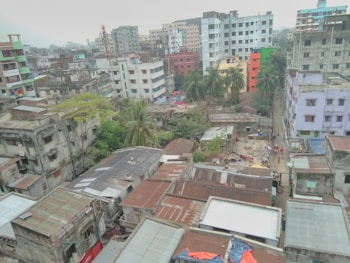 An urban skyline with a mix of residential and commercial buildings in varying states of repair. Some buildings are brightly colored, while others have a more aged appearance. A small number of trees and greenery are interspersed among the buildings, suggesting a densely populated area. Rooftops show a variety of materials and some are used for drying clothes. The area appears to be bustling with activity, indicated by people and objects scattered around.