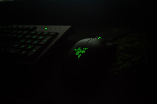 A dark-themed setup featuring a black computer mouse with a glowing green logo, placed next to a keyboard with illuminated green keys.