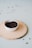 A white ceramic bowl filled with black peppercorns rests on a round wooden slice board. The background is a soft, pastel green with a few scattered peppercorns.