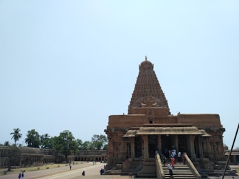 A large, ancient temple structure with intricate stone carvings and a towering central dome. The architecture reflects classical style with deep cultural significance. There are people walking around the temple and lush greenery surrounds the area, along with open courtyards and pathways.