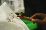 A close-up view of a person using a tool to apply wax on fabric for batik making. The hand holds a canting tool, which is releasing hot wax onto a white cloth with floral patterns drawn on it.