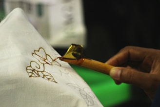 A close-up view of a person using a tool to apply wax on fabric for batik making. The hand holds a canting tool, which is releasing hot wax onto a white cloth with floral patterns drawn on it.