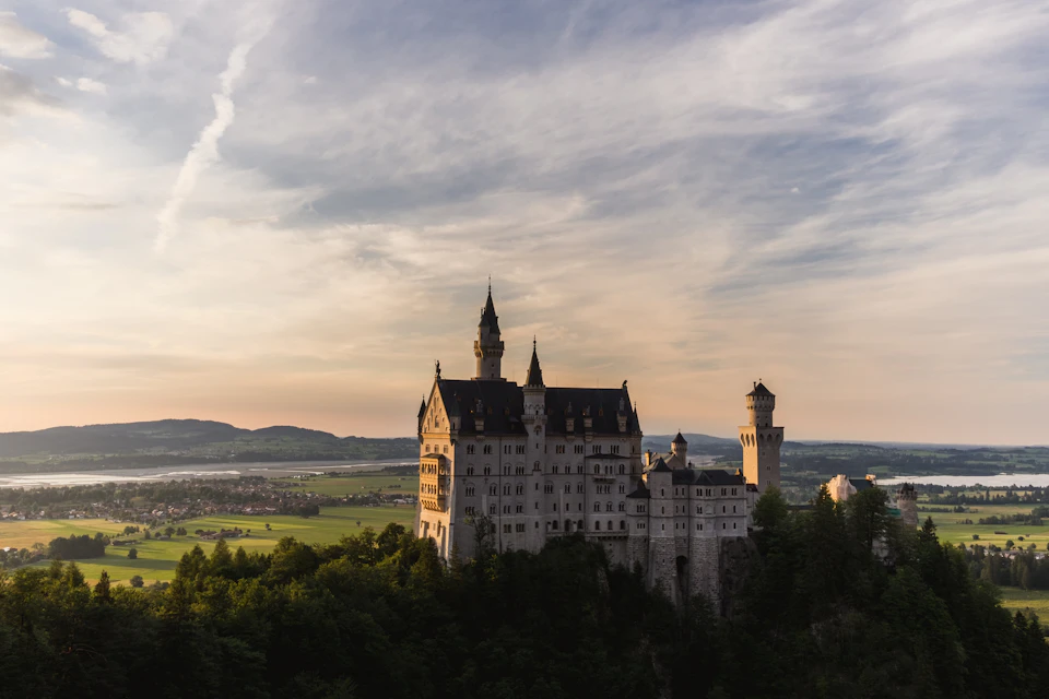 The image depicts a majestic castle perched atop a hill, surrounded by lush greenery and overlooking a serene valley below. The castle features multiple towers and spires that reach towards the sky, creating an imposing presence against the backdrop of the landscape. The perspective of the photo is from a high vantage point, providing a comprehensive view of the castle's grandeur and its harmonious integration with nature.

The colors in the image are predominantly green and brown, reflecting the natural surroundings and adding to the overall tranquility of the scene. The sky above is filled with clouds, suggesting that it might be either early morning or late afternoon when the sun casts a soft light over the landscape.